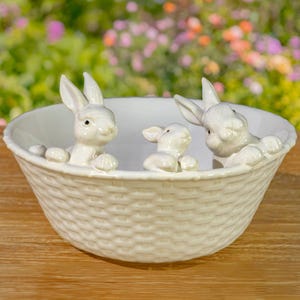 May include: A white ceramic bowl with a woven basket design. Three white bunny figurines peek over the rim. The bowl is set on a wooden surface with a blurred floral background, perfect for Easter decor.
