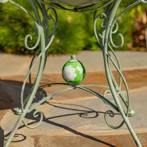 Puede incluir: Un soporte de metal verde claro decorativo para plantas con un adorno en forma de globo. El adorno es verde con detalles blancos, que se asemeja a un mapa. El soporte tiene un dise&ntilde;o enrollado y est&aacute; colocado sobre una superficie de piedra.
