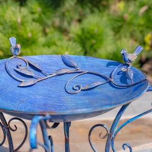 Puede incluir: Un bebedero para p&aacute;jaros de metal azul con detalles decorativos de hojas y vides. Dos peque&ntilde;os p&aacute;jaros azules se posan en el borde. El bebedero tiene una superficie texturizada y patas ornamentadas y curvas.