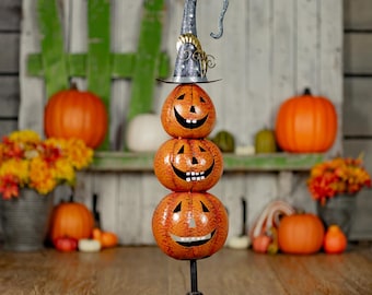 Puede incluir: Una pila vertical de tres calabazas de metal naranja sobre una base de pedestal negra. Cada calabaza presenta ojos recortados y una sonrisa dentada. La calabaza superior lleva un sombrero de bruja de metal azul oscuro alto y curvado.