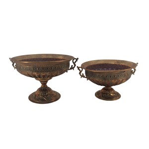 May include: Two antique bronze-colored decorative bowls. The bowls have ornate detailing around the rims and bases, with handles on either side. The interior of the bowls has a dark, textured surface. The bowls are of different sizes.