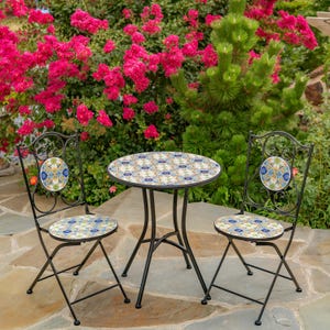 May include: A black metal bistro set with a round table and two chairs. The table and chair seats feature a colorful tile mosaic design. The chairs have decorative backrests with matching mosaic accents. The set is outdoors, with a backdrop of pink flowers and greenery.