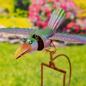 May include: A colorful metal bird with a long beak and large eyes is perched on a metal stand. The bird's wings are spread out and it appears to be in flight. The bird is painted in shades of blue, green, purple, and orange.