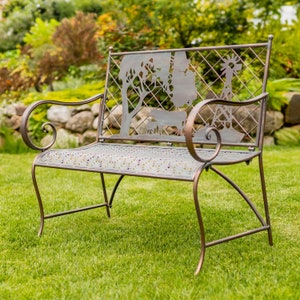 May include: A metal garden bench with a decorative cut-out design featuring a windmill, trees, and cows. The bench is brown and has a rustic look.