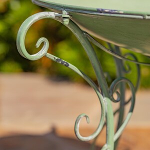 Puede incluir: Primer plano de una mesa de metal verde claro desgastado con un dise&ntilde;o de pata en espiral. La mesa tiene una parte superior redonda y un acabado envejecido, lo que sugiere un estilo antiguo o vintage. El fondo est&aacute; borroso.
