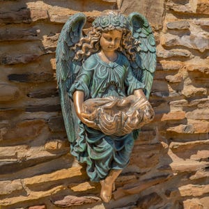 May include: A wall-mounted angel sculpture with a verdigris and bronze finish. The figure features large feathered wings, a floral crown, and flowing robes, while holding a large, open seashell basin in both hands with a downward gaze.