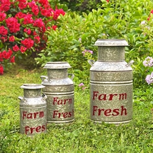 May include: Three galvanized metal milk cans of varying sizes are displayed outdoors in a grassy area. The cans have a rustic, textured finish and the words "Farm Fresh" are painted in red. The background features green foliage and red flowers.