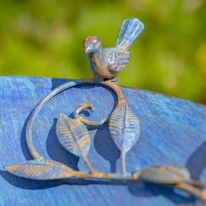 Puede incluir: Primer plano de un p&aacute;jaro de metal azul posado en una rama decorativa con hojas. El p&aacute;jaro y la rama tienen un aspecto envejecido y desgastado con toques de marr&oacute;n y naranja. El fondo es verde borroso.