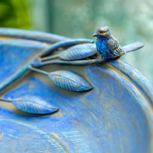 May include: Close-up of a blue birdbath with a small bird figurine perched on the edge. The birdbath has a distressed, aged appearance with blue and gold tones. Decorative leaves are sculpted around the rim, adding to the garden theme.