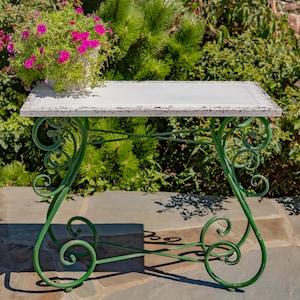 May include: A rectangular console table with a distressed white top and a green metal base featuring scrollwork legs. The table has a flat surface and a cross-brace support structure. A white pot filled with bright pink flowers sits on the table.
