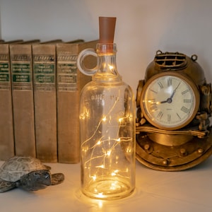 May include: A clear glass bottle with a brown cork and a handle, filled with small, warm white LED lights. The bottle is labeled "ONE PINT". A vintage-style clock and books are in the background.