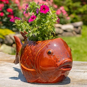 May include: A ceramic fish-shaped planter in shades of brown, filled with vibrant green foliage and bright pink flowers. The planter has a glossy finish and is placed outdoors.
