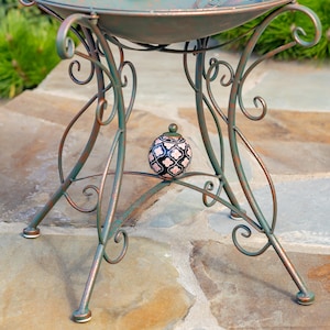 Puede incluir: Una mesa de metal decorativa para exteriores con un dise&ntilde;o de volutas en tonos verdes y &oacute;xido. La mesa tiene una parte superior redonda y patas curvas. Un peque&ntilde;o tarro de cer&aacute;mica estampado con tapa se encuentra debajo.