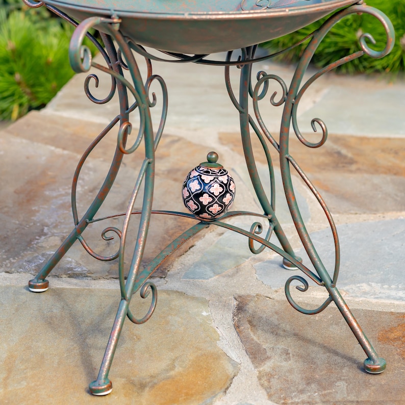 Puede incluir: Una mesa de metal decorativa para exteriores con un dise&ntilde;o de volutas en tonos verdes y &oacute;xido. La mesa tiene una parte superior redonda y patas curvas. Un peque&ntilde;o tarro de cer&aacute;mica estampado con tapa se encuentra debajo.