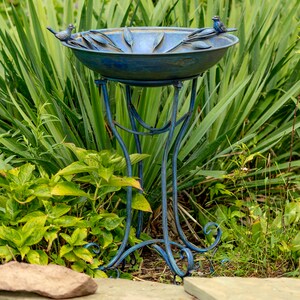 May include: A blue metal bird bath with a decorative design, featuring two small bird figurines perched on the rim. The bird bath is supported by a slender, ornate stand. The bird bath is surrounded by lush green foliage.