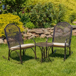 May include: Two dark bronze metal rocking chairs with arched backs featuring a circular lattice pattern and beige seat cushions, accompanied by a matching round side table with a decorative metal base and a circular lattice top surface.