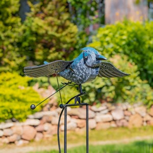 May include: A blue and gray metal bird wind spinner with black metal wings and a black metal stand. The bird is perched on a black metal rod that is attached to the stand. The wind spinner is in a garden setting.