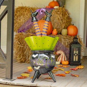 May include: A Halloween decoration featuring a cauldron with the word "Bewitched" in green lettering. The cauldron has three black legs and a lime green bowl with black polka dots. Witch legs with orange and purple striped stockings emerge from the bowl.
