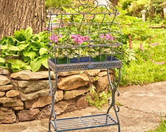 May include: An ornate, wrought-iron plant stand with a cage-like top, holding several potted pink flowers. The stand has a distressed, verdigris finish and sits on a stone patio in a garden setting. The design includes scrollwork and a fleur-de-lis finial.