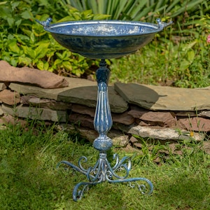 May include: A blue birdbath with a distressed finish, featuring a wide circular basin with two small bird figures on the rim, supported by a fluted pedestal and a decorative base with four curved, scroll-like metal legs for stability.