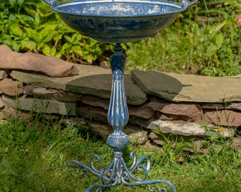 May include: A distressed blue metal bird bath with a round basin and a decorative pedestal base. Two small bird figurines perch on the rim. The bird bath stands on a grassy lawn, with a stone wall and greenery in the background.