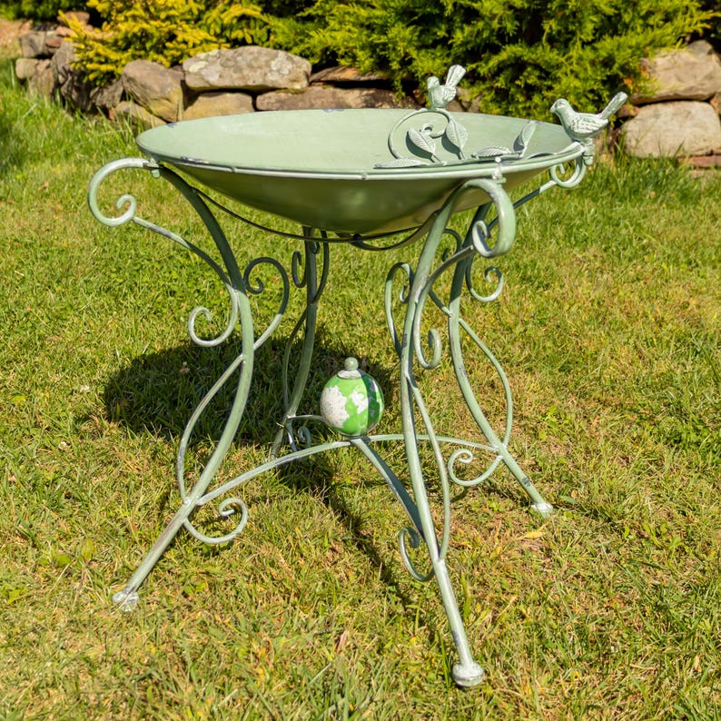 Puede incluir: Un bebedero para p&aacute;jaros de metal verde salvia con una base decorativa con volutas. El cuenco tiene un borde festoneado y est&aacute; adornado con tres peque&ntilde;as figuras de p&aacute;jaros. Un adorno en forma de globo est&aacute; centrado en la base. El bebedero est&aacute; al aire libre sobre un c&eacute;sped.