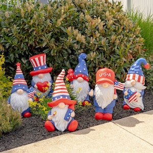 May include: Six patriotic gnomes with red, white, and blue hats and outfits. The gnomes are standing on a bed of mulch and are surrounded by green plants.