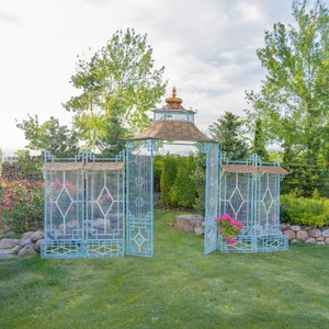 May include: A blue metal garden archway with a gold roof and decorative latticework. The archway is surrounded by green foliage and flowers.