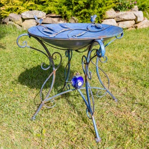 Puede incluir: Un bebedero para p&aacute;jaros de metal azul con un dise&ntilde;o decorativo. El bebedero tiene una cuenca redonda con detalles de hojas y p&aacute;jaros, sostenida por un marco de metal enrollado. Una peque&ntilde;a bola decorativa azul y blanca est&aacute; unida al marco.