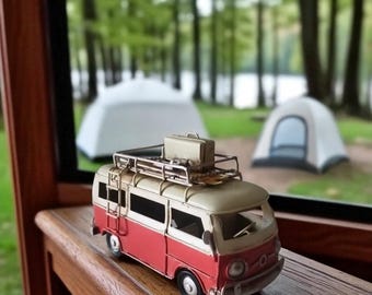 May include: A miniature red and cream-colored camper van with a luggage rack and suitcase on top. The van is parked on a wooden surface, with a blurred background of tents and trees, suggesting a camping theme.