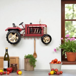 May include: A red metal wall-mounted tractor-shaped rack featuring the text OLD TRACTOR on the side. The tractor has two large black wheels with cream-colored rims and includes several small hooks underneath for hanging items on the wall.