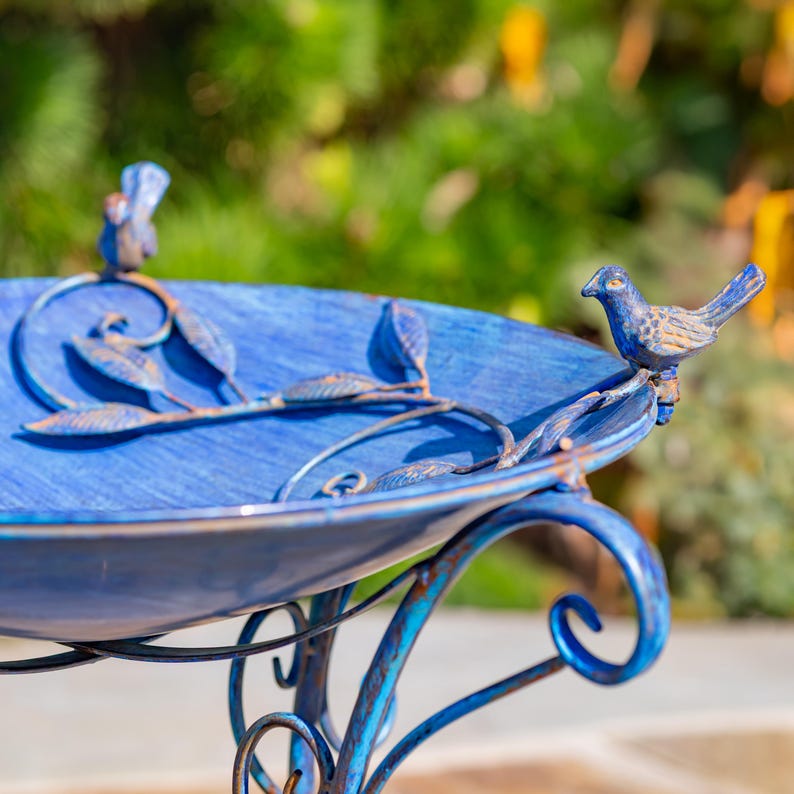 Puede incluir: Un bebedero para p&aacute;jaros de metal azul con un dise&ntilde;o decorativo. El bebedero tiene una cuenca redonda con una superficie texturizada y est&aacute; sostenido por un soporte de metal curvado. Dos peque&ntilde;as figuras de p&aacute;jaros azules est&aacute;n posadas en el borde de la cuenca.