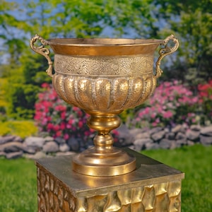 May include: A metallic gold-toned urn with a wide, flared rim and two ornate, curved handles. The upper section features a detailed, textured band with circular patterns, while the lower body has vertical, hammered fluting above a pedestal base.