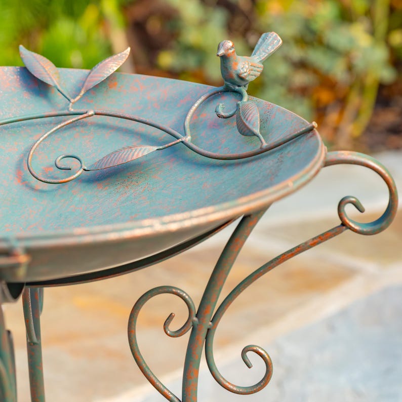 Puede incluir: Un bebedero decorativo para p&aacute;jaros con un acabado desgastado de color verd&iacute;n. El bebedero de metal presenta un peque&ntilde;o p&aacute;jaro posado en el borde, con detalles de hojas y vides. El bebedero tiene una cuenca redonda y soportes decorativos en espiral.