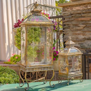 May include: Two ornate, antique-style lanterns with glass panels and decorative metal frames. The larger lantern stands on a scrolled base. The lanterns have a gold finish and are displayed outdoors near greenery and flowers.