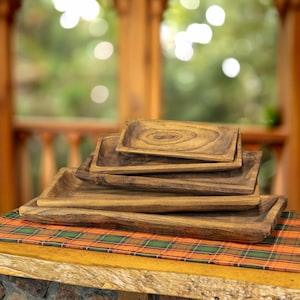 May include: Five rectangular wooden serving trays of varying sizes, stacked on a table. The trays have a natural wood grain finish. They are displayed on a table with a plaid runner, with a blurred outdoor background.