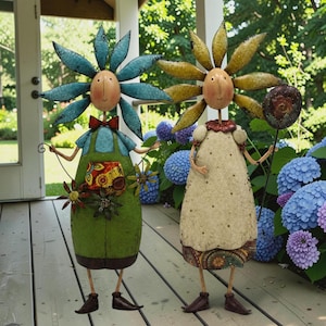 May include: Two metal garden sculptures featuring oval faces and petal-shaped headpieces. The left figure wears a green apron with a patterned pocket and blue petals. The right figure wears a cream dress with a patterned hem and yellow petals.