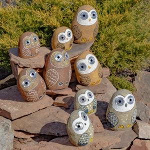 May include: A selection of stone owl ornaments in varying sizes and shades of brown, tan, and grey. Each owl features large, round eyes and decorative wing patterns. The owls are displayed on a bed of rocks with green foliage in the background.