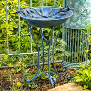 May include: A blue metal bird bath with a decorative leaf and bird design. The bird bath has a round basin and is supported by a three-legged stand. The bird bath is placed in a garden setting with green foliage and a decorative fence in the background.