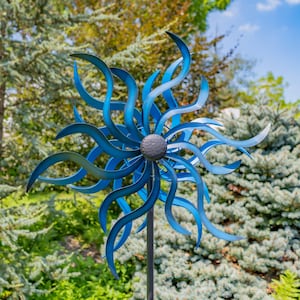 May include: A metal wind spinner featuring multiple curved, wave-like blades in varying shades of blue, radiating from a central dark gray textured circular hub, mounted on a thin, straight, dark gray metal pole.