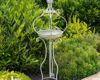 May include: A decorative, antique-style metal plant stand with a distressed, light green finish. The stand features a circular basin and ornate, curved supports. The top has a decorative finial with a blue and white floral design. The stand is surrounded by lush greenery.