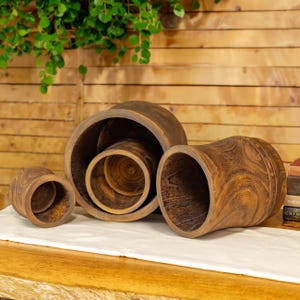 May include: A collection of four wooden planters in varying sizes, arranged on a white cloth. The planters are cylindrical with a rich, dark brown finish, showcasing the natural wood grain. The largest planter is on its side, revealing its interior.