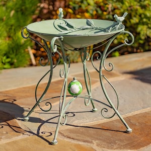 Puede incluir: Un bebedero decorativo de metal verde salvia con un dise&ntilde;o detallado. La cuenca en forma de cuenco presenta detalles de hojas y p&aacute;jaros, sostenida por patas curvas y ornamentadas. Un peque&ntilde;o globo decorativo est&aacute; centrado en las patas.