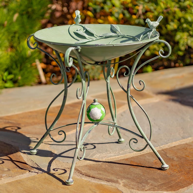 Puede incluir: Un bebedero decorativo de metal verde salvia con un dise&ntilde;o detallado. La cuenca en forma de cuenco presenta detalles de hojas y p&aacute;jaros, sostenida por patas curvas y ornamentadas. Un peque&ntilde;o globo decorativo est&aacute; centrado en las patas.