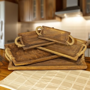 May include: Four wooden serving trays of different sizes, stacked together. Each tray is rectangular with rope handles and a dark brown finish. The trays are designed for serving food or displaying items.