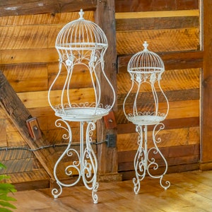 May include: Two white metal birdcage-style plant stands with dome-shaped tops, curved wire frames, and decorative floral accents. Each stand features a circular base platform supported by ornate, scrolled legs, creating a tall, open structure.