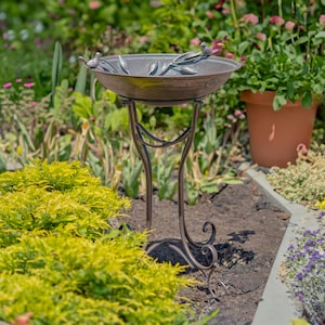 May include: A bronze-colored bird bath with a decorative design. The bowl has leaf and bird accents. The bird bath is supported by a curved metal stand, and is surrounded by greenery and flowers.