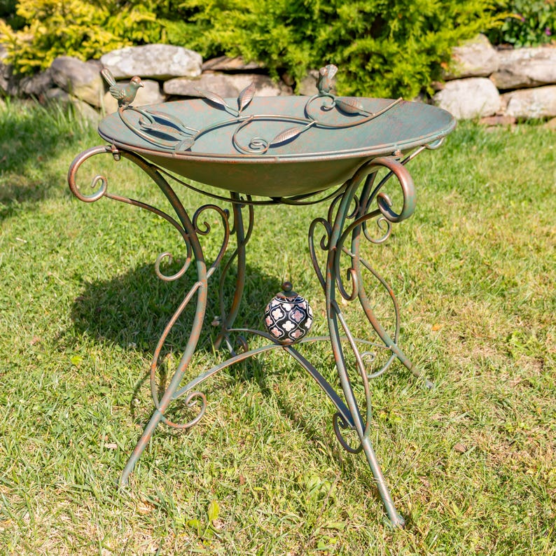 Puede incluir: Un bebedero decorativo de metal con un acabado verde desgastado. El cuenco est&aacute; adornado con detalles de hojas y p&aacute;jaros. Las patas ornamentadas presentan volutas y sostienen un globo decorativo de cristal. El bebedero est&aacute; sobre c&eacute;sped.