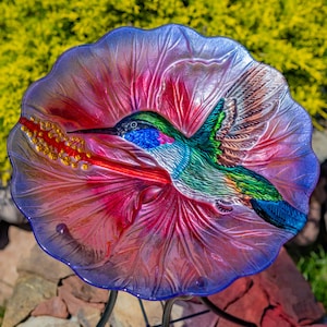 May include: A decorative glass hummingbird feeder with a vibrant design. The feeder features a colorful hummingbird in flight, set against a red and pink flower with a blue rim. The feeder is supported by a black metal stand.