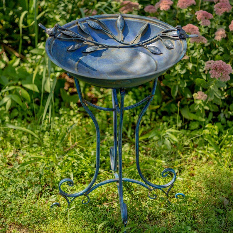 May include: A blue metal bird bath with a decorative leaf and bird design. The bird bath has a round basin and a three-legged stand. The stand has decorative scrollwork at the base. The bird bath is outdoors in a garden setting.